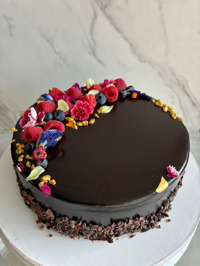 Custom cakes Oahu by Italian pastry chef. This cake is a chocolate sponge cake with chocolate pastry cream filling and a chocolate glaze finish, decorated with fresh berries, edible flowers, and gold leaf. 