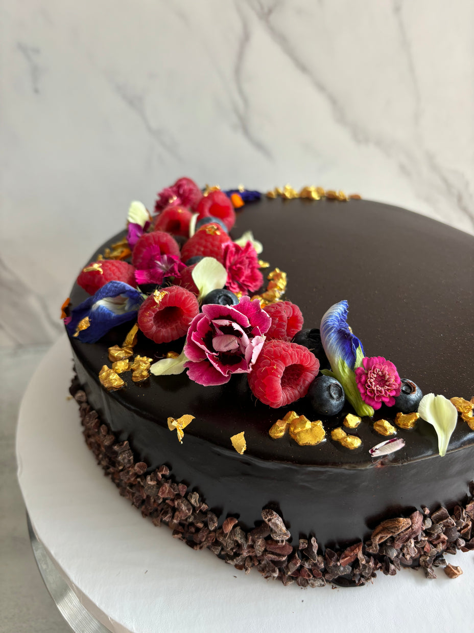 Custom cakes Oahu by Italian pastry chef. This cake is a chocolate sponge cake with chocolate pastry cream filling and a chocolate glaze finish, decorated with fresh berries, edible flowers, and gold leaf.