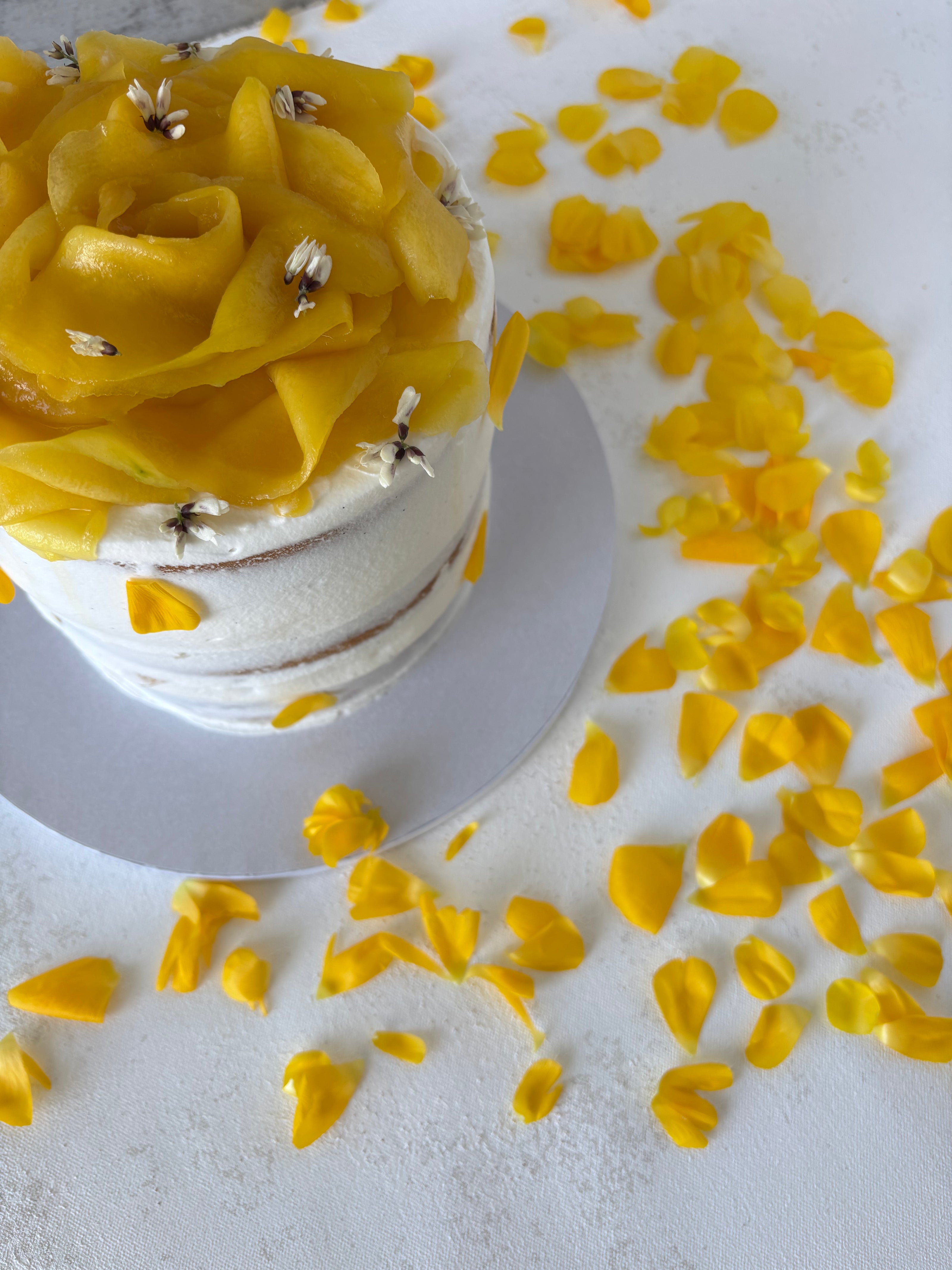 This is a round vanilla and mango cake. It is made of vanilla sponge, white chocolate chantilly, and fresh mango bites. It is decorated with a flower made of mango slices and fresh edible flowers.