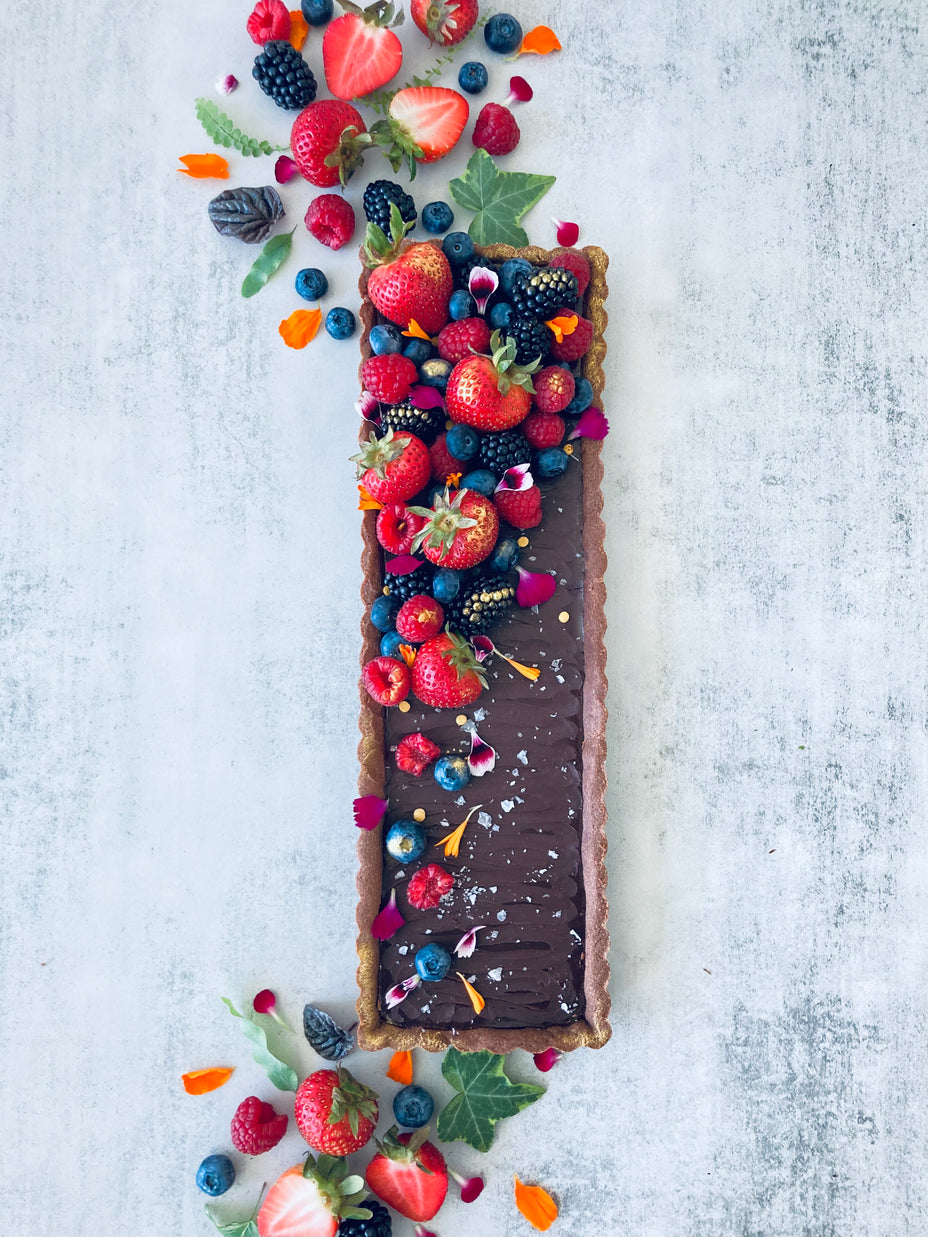 A rectangular chocolate tart with a buttery chocolate shell and rich chocolate ganache filling, decorated with fresh mixed berries, edible flowers, and Maldon salt flakes. 