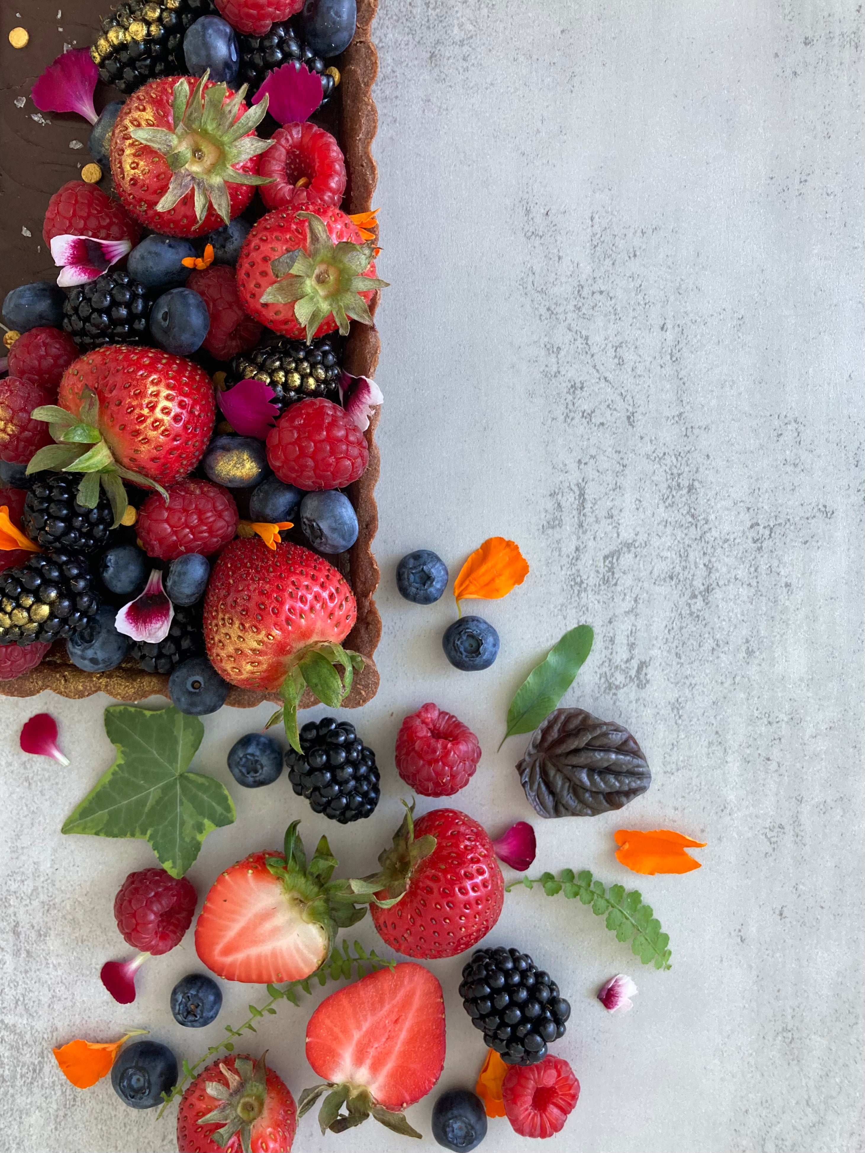 A rectangular chocolate tart with a buttery chocolate shell and rich chocolate ganache filling, decorated with fresh mixed berries, edible flowers, and Maldon salt flakes. 
