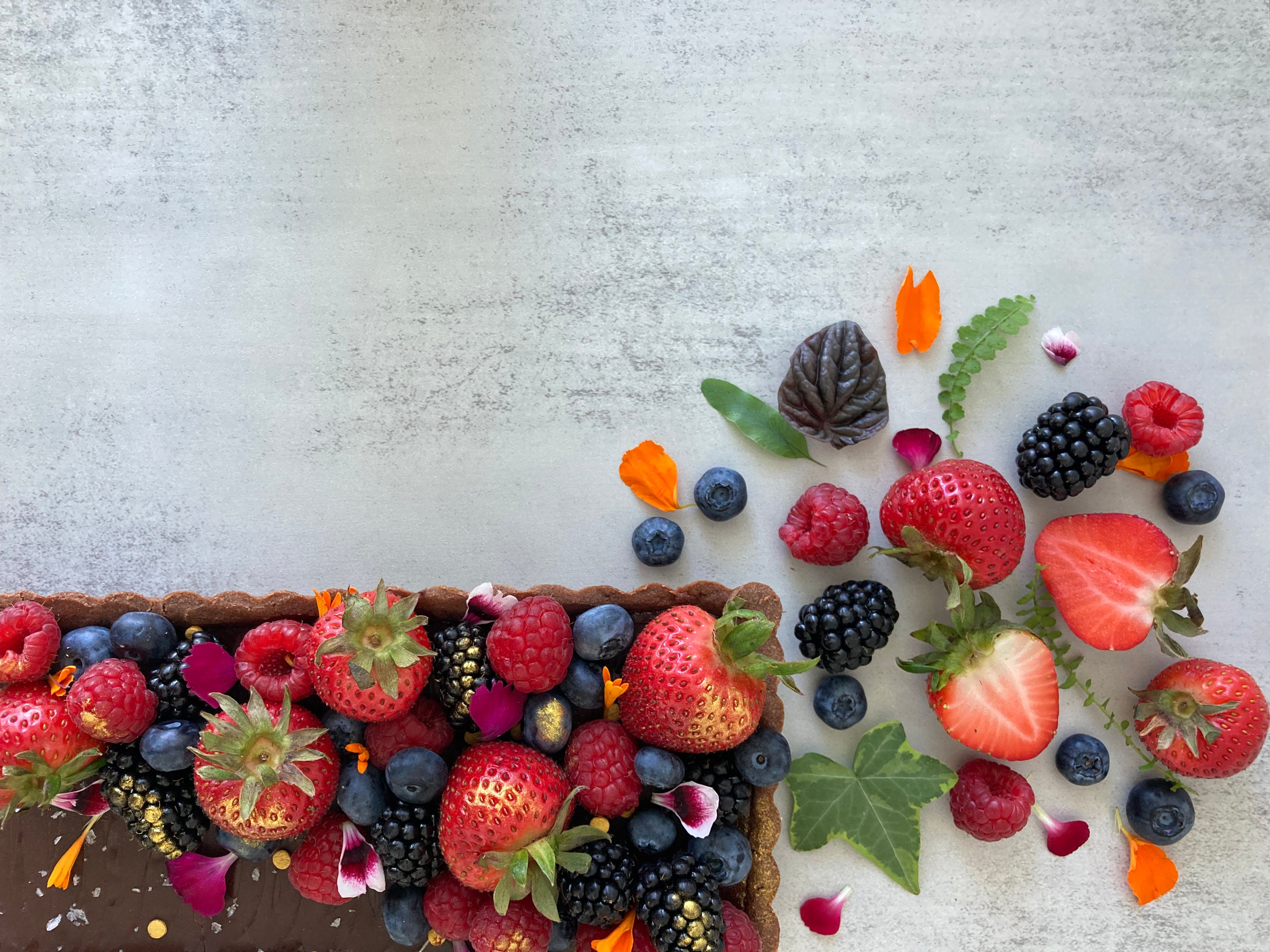 A rectangular chocolate tart with a buttery chocolate shell and rich chocolate ganache filling, decorated with fresh mixed berries, edible flowers, and Maldon salt flakes. 